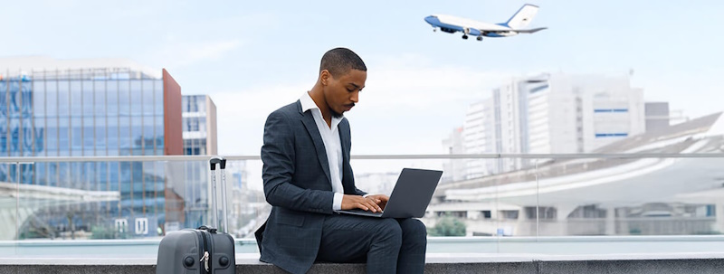 A businessman working on a laptop at the airport, with a suitcase beside him and an airplane flying overhead.