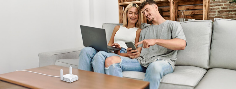 A young couple sitting on a sofa at home, using a laptop and smartphone together, with the ASUS RT-BE58 Go on the table in front of them.